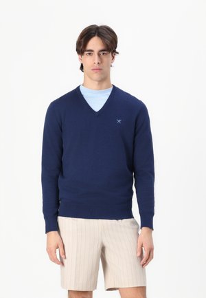 Granatowy sweter z dekoltem w serek, wykonany z miękkiego materiału, z prążkowanymi mankietami i dołem, noszony na jasnoniebieską koszulę, w połączeniu z beżowymi szortami w paski.