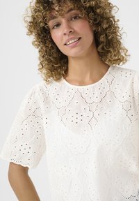 Blusa bianca ricamata con un motivo floreale e dettagli a taglio, scollo rotondo e maniche corte a sbuffo. Tessuto in cotone con una consistenza morbida.