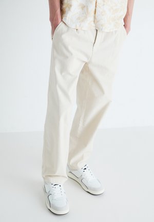 Helle Baumwollhosen mit lockerem Schnitt, ausgestattet mit Seitentaschen und einer glatten Textur, kombiniert mit weißen Sneakers.