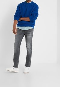 Suéter de punto azul, camisa azul claro, vaqueros grises y zapatos blancos sin cordones. El suéter tiene un cuello redondo y puños acanalados; los vaqueros presentan un patrón desgastado.