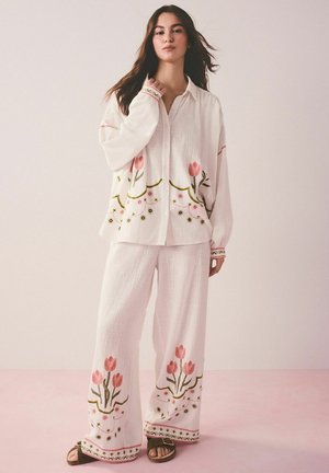 Woman wearing loose white button-up shirt and wide pants, both with pink floral embroidery, standing on pink floor against light wall.