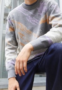 Personne portant un pull gris avec des motifs abstraits orange et violet, associé à un pantalon bleu foncé, assise à l'extérieur.