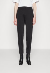 Černé látkové kalhoty s úzkým střihem, které mají hladkou texturu, jemné přední sklady a středně vysoký pas. V páru s černými kotníkovými botami.