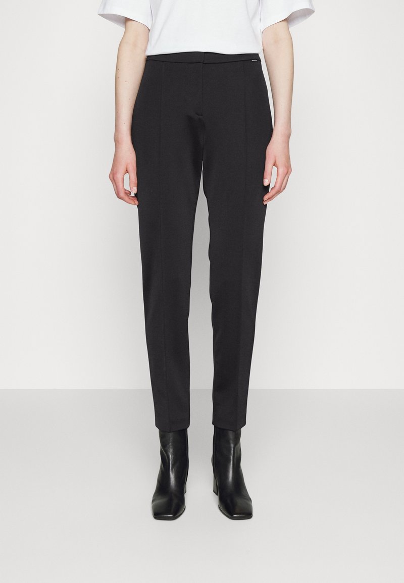 Černé látkové kalhoty s úzkým střihem, které mají hladkou texturu, jemné přední sklady a středně vysoký pas. V páru s černými kotníkovými botami.