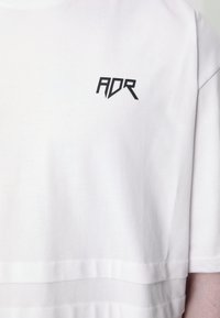 White cotton T-shirt with short sleeves, featuring a black "ADR" logo near the neckline and a layered hem design. Smooth texture.