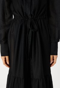 Musta mekko, jossa on teksturoitu, raidallinen kuviointi, vyötärölle kiristetty malli ja nauhatyyny. Pitkät hihat, joissa on pistekuvioinen kangas.