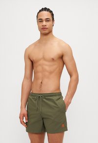 Shorts de baie verde măsliniu cu talie ajustabilă, având un detaliu cu logo portocalie în partea stângă jos. Fabricate dintr-un material ușor și neted.