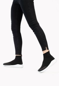 Czarne dzianinowe sneakersy za kostkę mają zamek błyskawiczny i białą gumową podeszwę. W połączeniu z dopasowanymi czarnymi dżinsami, podkreślającymi smukłą sylwetkę.