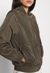 Olivově zelená bomber bunda vyrobená z lesklého nylonu, funkcí předního zipu, bočními kapsami, ribbované manžety a volným střihem.