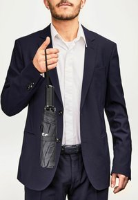 Parapluie noir plié tenu dans la main d'un homme, avec un élégant manche noir arborant le logo "BOSS", présenté contre un costume bleu marine.