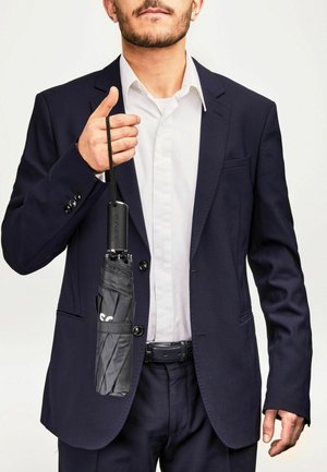 Parapluie noir plié tenu dans la main d'un homme, avec un élégant manche noir arborant le logo "BOSS", présenté contre un costume bleu marine.