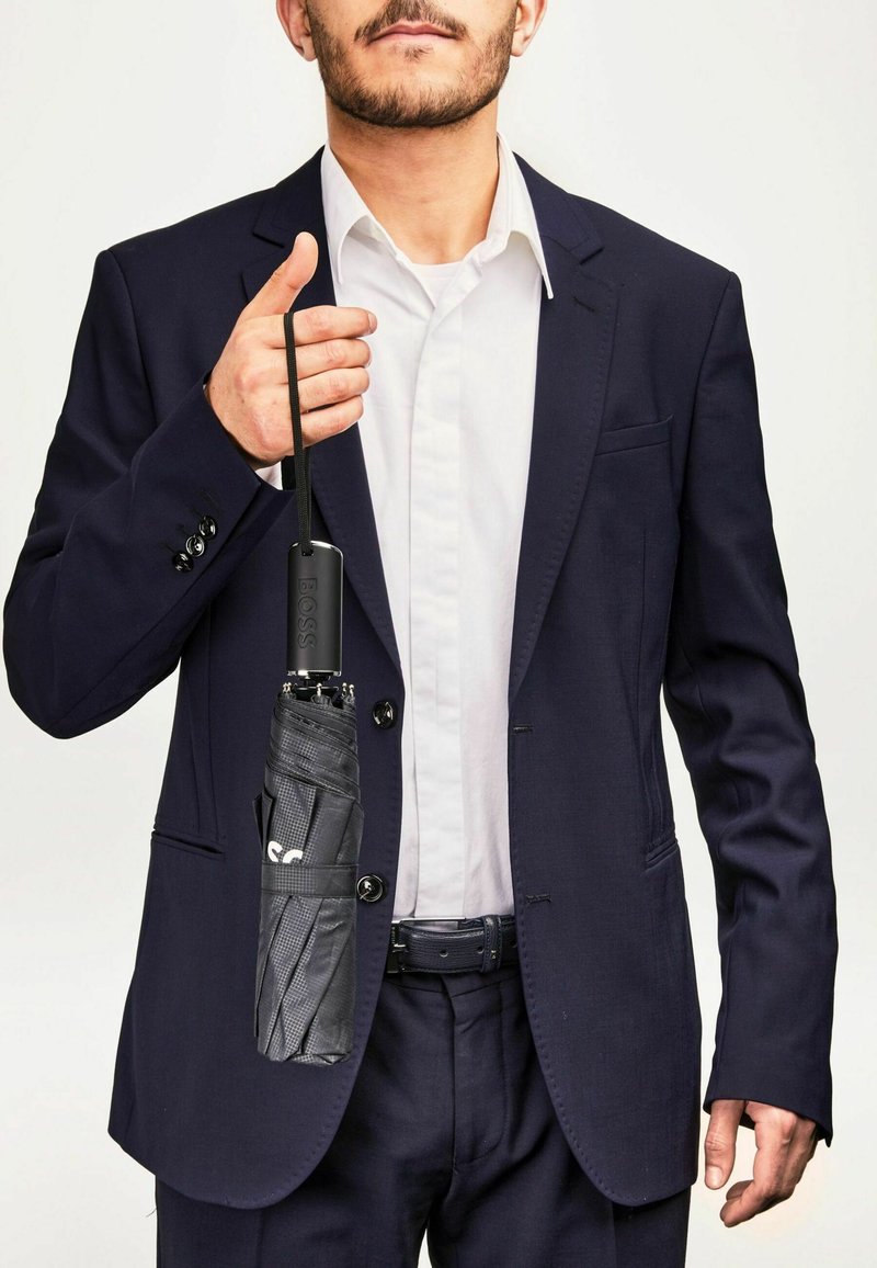 Parapluie noir plié tenu dans la main d'un homme, avec un élégant manche noir arborant le logo "BOSS", présenté contre un costume bleu marine.