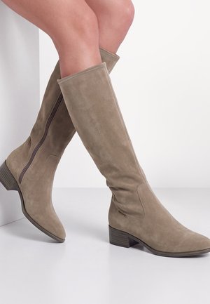 Bottes en daim taupe montant jusqu’au genou avec talons empilés bas et fermetures éclair latérales, portées sur des jambes croisées sur un fond blanc uni.