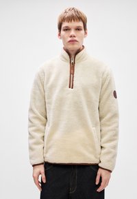 Fleece pullover i creme med halv lynlås, brune detaljer og sidelommer. Blød tekstur; har et rundt mærke på venstre ærme.