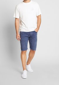 Bílý bavlněný tričko s krátkým rukávem a malou grafikou. Modré denimové šortky, po kolena, nošené s bílými teniskami. Neutrální pozadí.