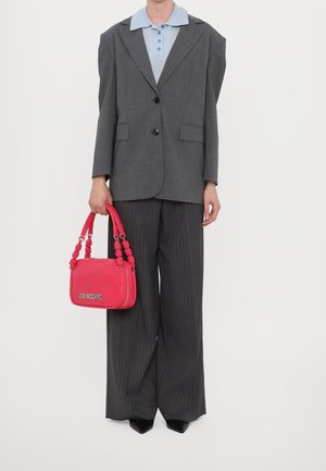 Person trägt einen übergroßen dunkel gestreiften Anzug und ein hellblaues Hemd mit Kragen, hält eine leuchtend pinke Love Moschino Handtasche seitlich.