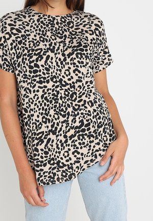 Kobieta w beżowej bluzce z krótkim rękawem w czarny wzór zwierzęcy oraz jasnoniebieskich dżinsach, stojąca na tle białym.