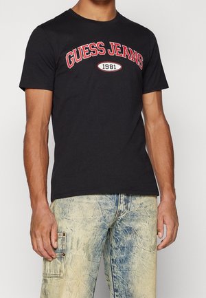 Mies pukeutunut mustaan "GUESS JEANS 1981" -t-paitaan ja vaaleansinisiin haalistuneisiin farkkuihin, joissa on taskut, seisoo yksiväristä taustaa vasten.