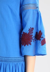 Blauwe katoenen blouse met geborduurde bordeauxrode bloemenontwerpen op de mouw, met kantdetails en een gatherd taille.