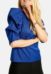 Žena s dlhými blond vlasmi, oblečená v modrom topu s balónovými rukávmi a čiernej sukni, usmieva sa s jednou rukou pri brade a druhou prekríženou.