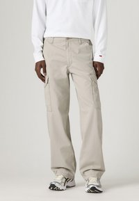Béžové cargo nohavice so bočnými vreckami, vo voľnom strihu a šitými detailmi, skombinované s bielou košeľou s dlhým rukávom a športovými topánkami.