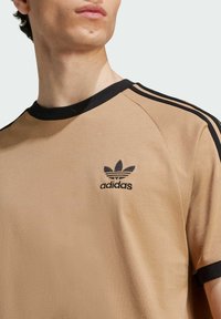 Camiseta de algodón color beige con cuello redondo de canalé negro, rayas laterales negras y un logo de Adidas bordado en negro en el pecho.