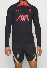 Pitkähihaista mustaa urheilupläkkäriä punaisilla yksityiskohdilla, jossa on "AXA"-logo ja Liverpool FC:n tunnus. Yhdistetty mustiin shortseihin ja Nike-brändäykseen.