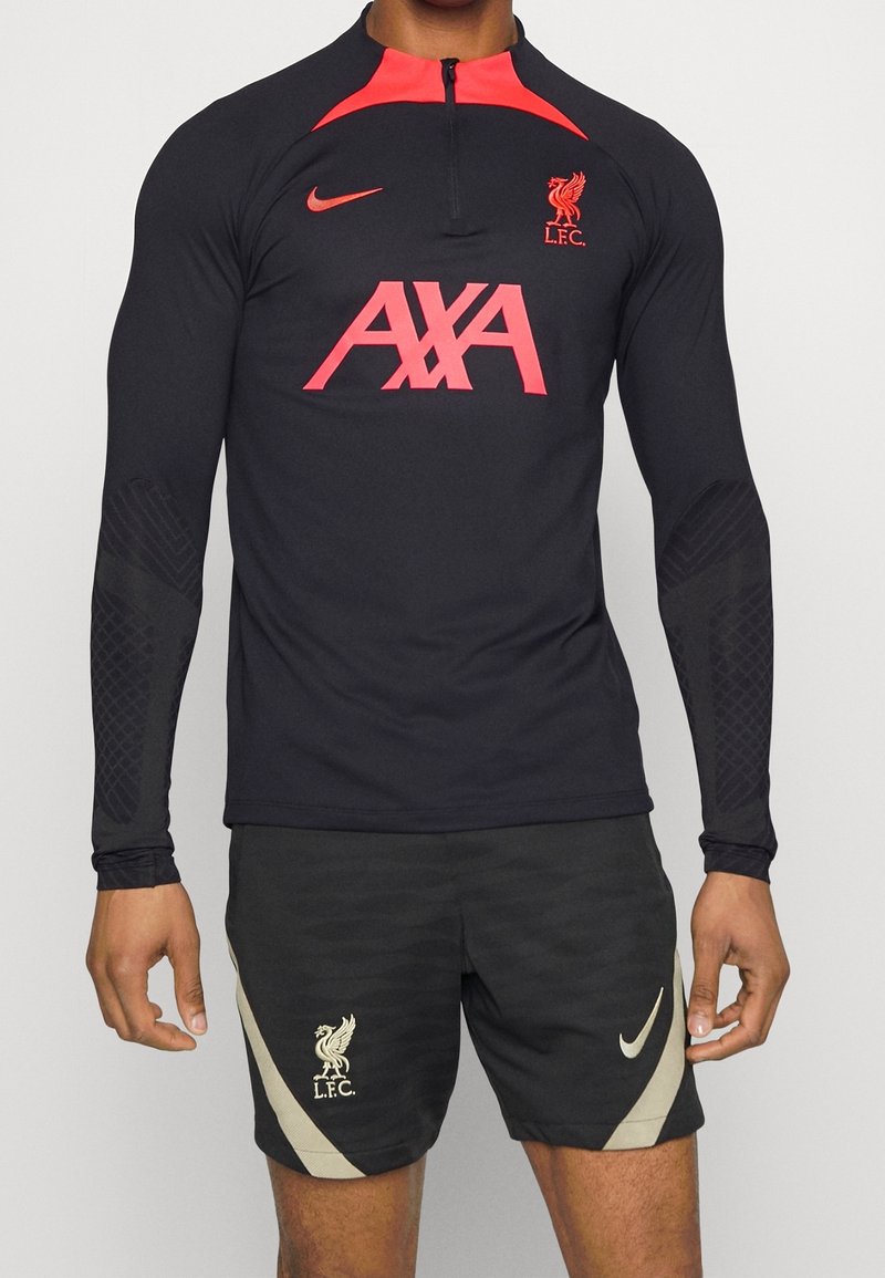 Pitkähihaista mustaa urheilupläkkäriä punaisilla yksityiskohdilla, jossa on "AXA"-logo ja Liverpool FC:n tunnus. Yhdistetty mustiin shortseihin ja Nike-brändäykseen.