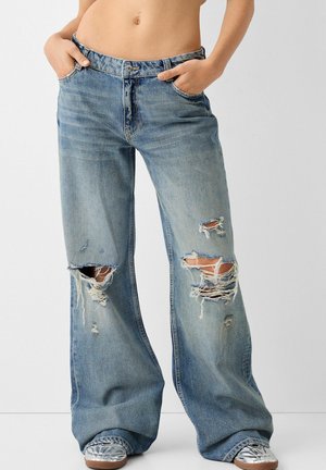 Person trägt weite, zerrissene blaue Jeans mit Händen in den Taschen und silberne Sneaker, steht vor schlichtem hellem Hintergrund.