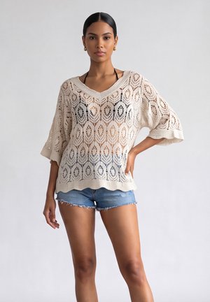 Vrouw draagt een beige gebreide haaktop over een zwarte bikini en blauwe denim shorts, staande met één hand op de heup tegen een effen achtergrond.