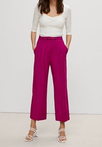 Fuksjowe spodnie o szerokich nogawkach z plisami z przodu, dwoma bocznymi kieszeniami i wysokim stanem, w połączeniu z dopasowaną białą bluzką i białymi sandałami na obcasie.