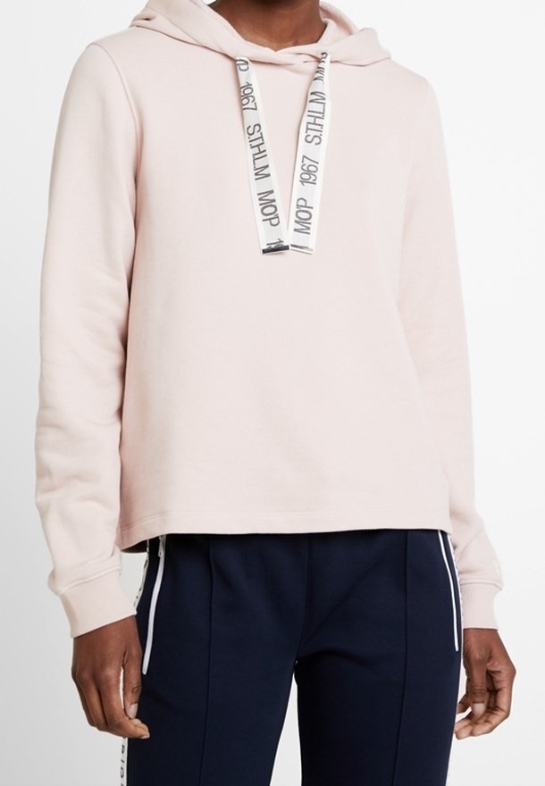 Sudadera con capucha rosa de tela suave, con un ajuste relajado, cordón con texto impreso y puños acanalados. Combinada con pantalones azul marino.