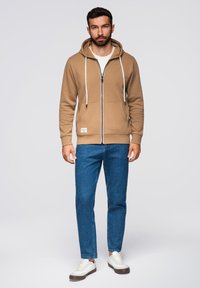 Felpa con zip color sabbia, cappuccio con cordino, tasche frontali e dettagli bianchi, abbinata a jeans blu e sneaker bianche.