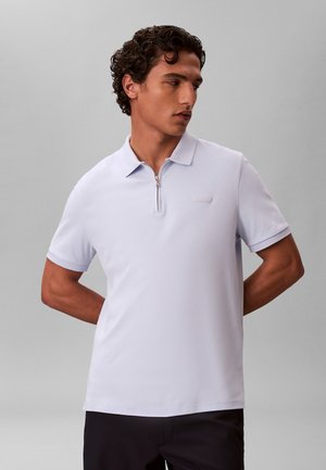 Homme aux cheveux bouclés portant un polo à manches courtes violet clair avec une fermeture éclair à l'avant, debout les mains derrière le dos.