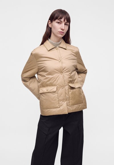 Femme portant une doudoune beige boutonnée avec poches à rabat et un pantalon large noir, debout les mains derrière le dos.