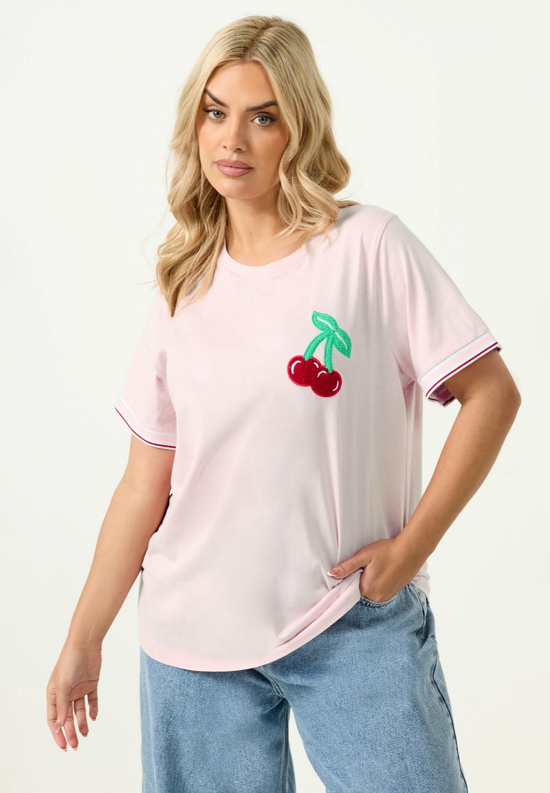 Femme blonde portant un t-shirt rose clair avec une broderie de cerisier sur la poitrine et un jean bleu, posant avec une main dans la poche.