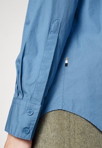 Ljusblå skjorta med knappar och tänkta ärmar, som har två blå knappar och en liten svart, vit och guldig etikett i kanten.