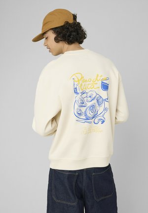 Personne portant un sweat beige avec une illustration de fruits de mer et un verre de vin dans le dos, associé à un jean foncé et une casquette marron.
