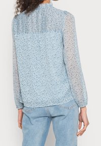 Blusa floreale azzurro chiaro con maniche lunghe trasparenti, colletto a volant e schiena arricciata. Presenta piccoli motivi floreali in diverse tonalità.