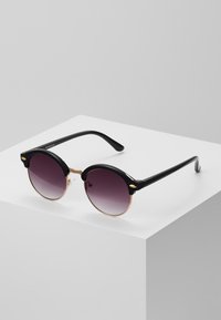 Gafas de sol redondas con montura de plástico negro y lentes morados degradados. Cuenta con acentos dorados y brazos de templado negro en ángulo.