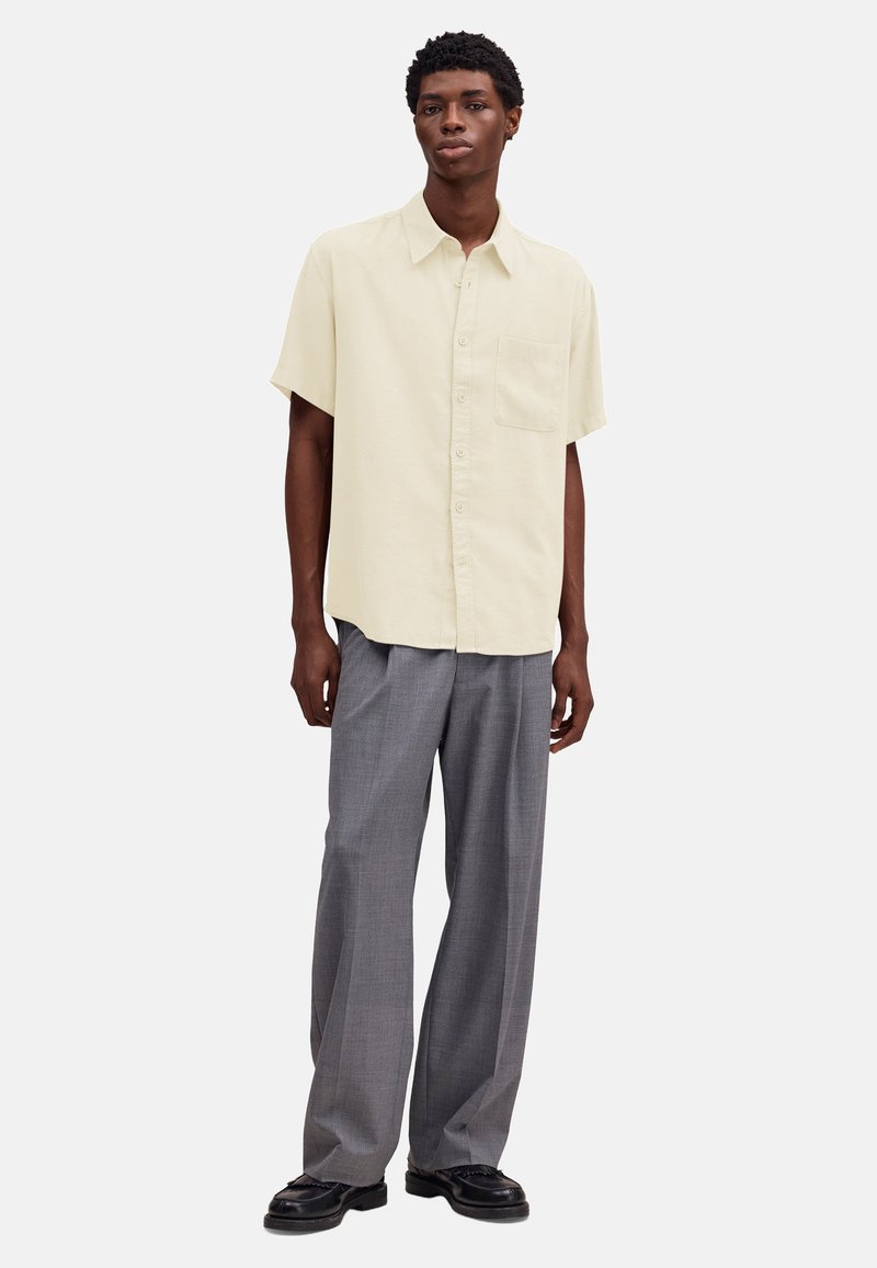 Homme portant une chemise beige à manches courtes avec des boutons, un pantalon gris à jambes larges et des chaussures en cuir noires, debout devant un fond uni.
