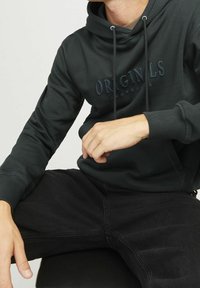 Ciemnozielona bluza z kapturem o teksturowanym wykończeniu, z centralnie umieszczonym haftowanym logo. W zestawie z czarnymi dżinsami, prezentującymi swobodny styl.