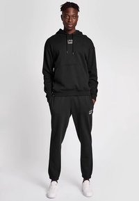 Svart hoodie- och mjukisbyxeoutfit med framficka, dragsko i huvan, elastiska muddar och logodetaljer. Vita sneakers kompletterar looken.