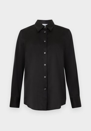 Femme portant une chemise noire ample boutonnée et un pantalon beige à jambes larges, debout devant un fond blanc uni.