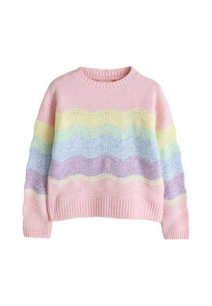 Young girl with curly hair wearing a pastel rainbow sweater, white wide-leg pants, and gold sneakers, standing on a beige background.