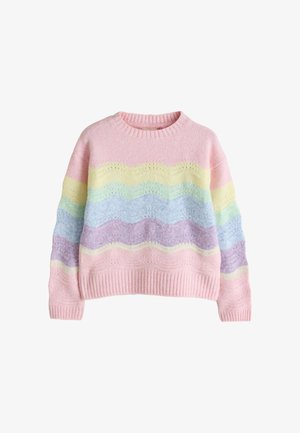 Junges Mädchen mit lockigem Haar, das einen pastellfarbenen Regenbogenpullover, weiße weite Hosen und goldene Sneakers trägt, steht vor einem beigen Hintergrund.
