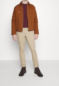 Braune Canvasjacke mit einer Brusttasche, kombiniert mit einem bordeauxfarbenen Pullover, beigen Hosen und dunkelbraunen Schnürstiefeln. Einfaches Design, taillierte Passform.