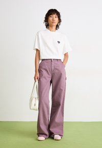 T-shirt blanc avec un logo de cœur noir, associé à un pantalon lavande taille haute et large. Les accessoires incluent un petit sac à main blanc et des chaussures rose pâle.