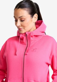Rosa zip-up hoodie i mjukt tyg, med huva, dragsko och en logotypdetalj på bröstet. Slät textur och avslappnad passform.