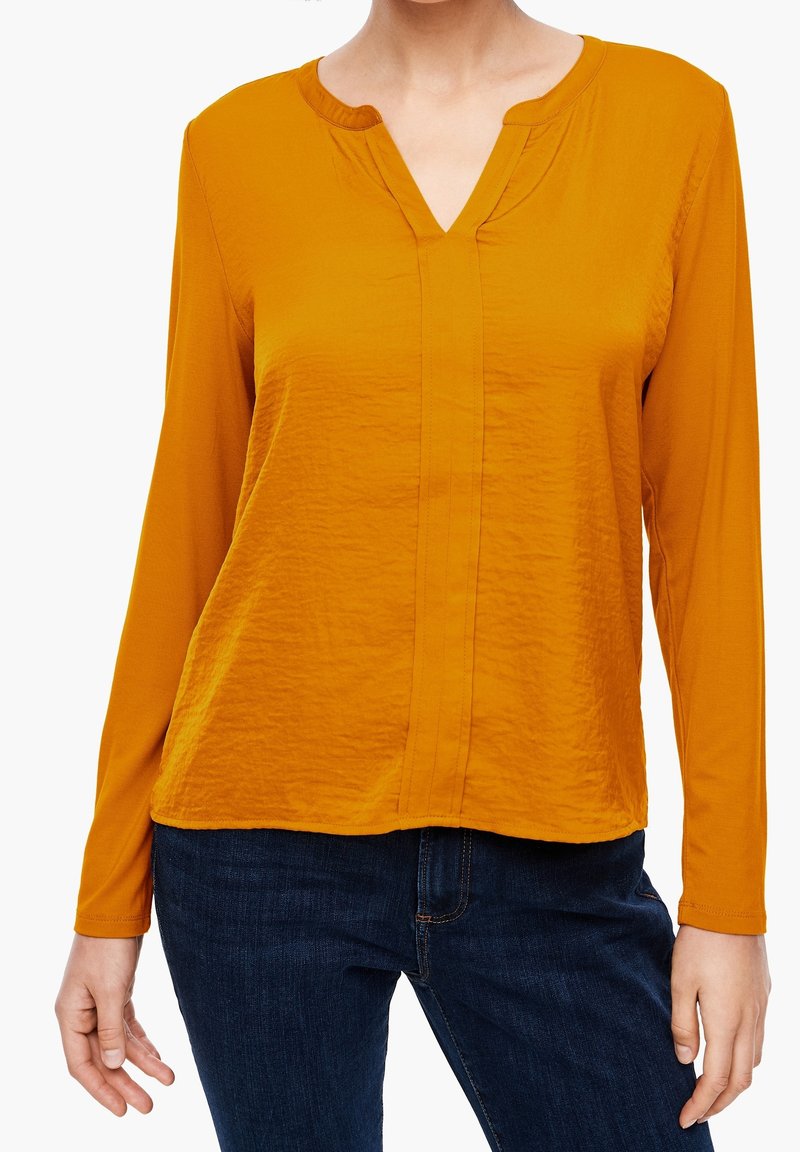 Chemise à manches longues jaune moutarde avec un col en V, en tissu texturé, dotée d'une couture centrale et d'une coupe décontractée, associée à un jean noir.